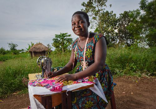 Ugandan woman smiling and looking at the camera, while she is sewing a piece of fabric