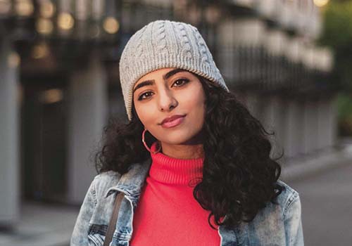 Portrait of Anahita Seraji. Anahita smiles at the camera, wears a knit grey hat on her beautiful black long curly hair. She's also wearing a red turtle neck sweater under a jeans jacket. 