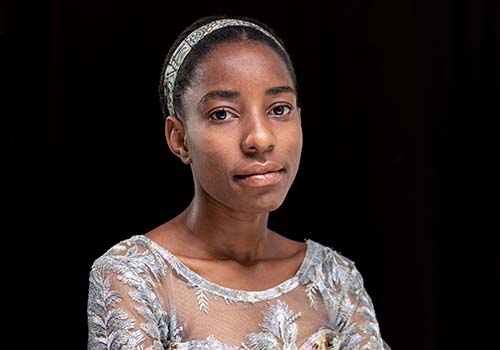 Portrait of Nadege Cibogo Felista wearing a white embroidery dress. Nadege has her hair up and is wearing a matching hair band. 