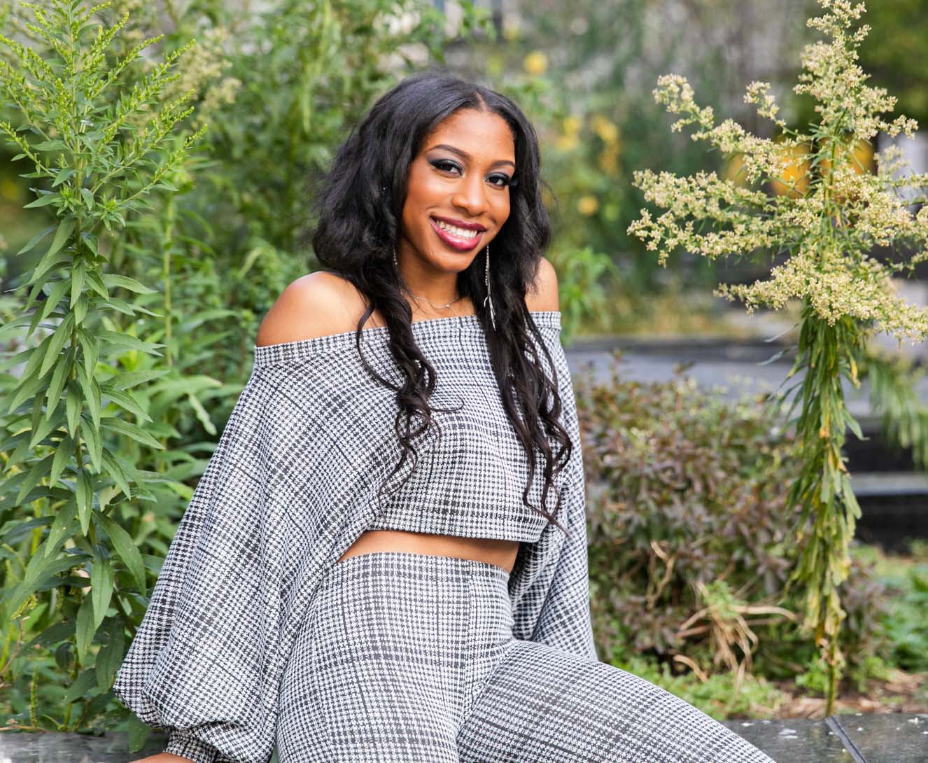 Young filmmaker Aria Harrell sitting outdoors and smiling to the camera. Aria wears a stripped black and white top and pant outfit and is surrounded by greenery.