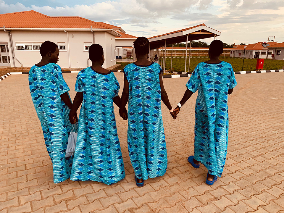 Four women at Terrewode hostpital in Uganda holding hands while they walk. They are all wearing the same turquoise blue gown.