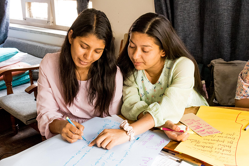 Two young women working together on a paper. One is pointing to the paper while the other one writes on it.