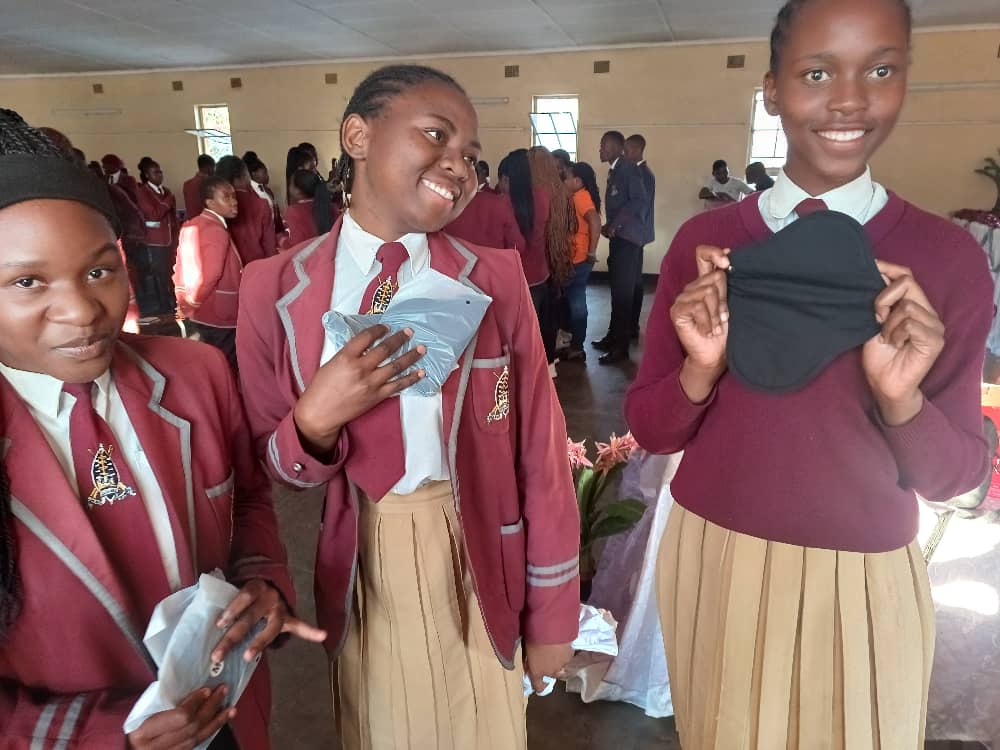 Image shows three Zimbabwean female students in school uniform showing period pads and smiling to the camera